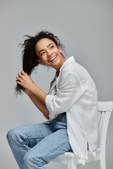 Obraz premium A young African American woman with curly hair sits on a white chair, smiling and playfully touching her hair.