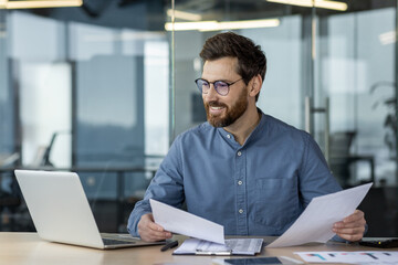 Fototapeta premium Smiling businessman working with documents and laptop in modern office