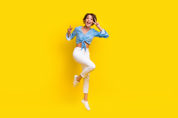 Full length photo of eccentric ecstatic lovely girl dressed blue top showing v-sign on eye jumping isolated on yellow color background