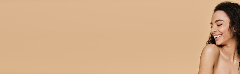 Obraz premium A beautiful young woman with natural makeup smiles brightly against a warm beige backdrop.