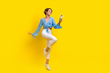 Full size photo of elegant adorable woman wear print blouse jumping read notification on smartphone isolated on yellow color background