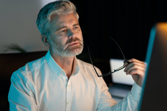 Thoughtful Senior Man Working Late Night In Office, Holding Glasses And Looking At Computer