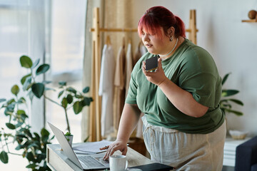 A woman stands at a table, laptop open, phone in hand, looking thoughtful.