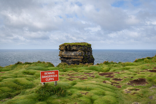 a red beware sign on the edge of a grassy cliff in Ireland with a sea stack rock formation