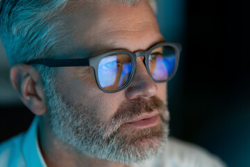 Close-Up Of Mature Man With Glasses Focused On Computer Screen, Reflections In Lenses, Dim Lighting, Late Night Work, Technology And Concentration © puhhha