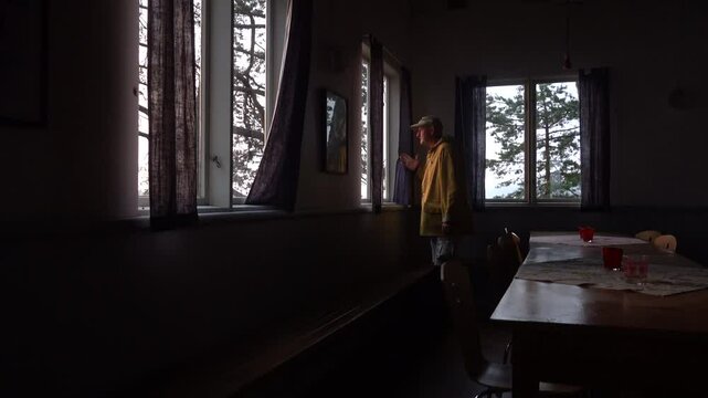 Stockholm, Sweden A man with a yellow raincoat looks out the window at dawn in a small cabin by a lake. 