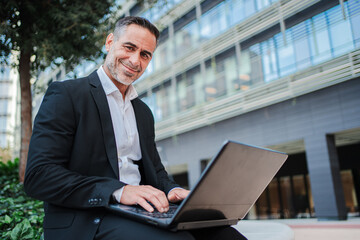 Individual portrait of a mature businessman sitting outside in the financial district, working with...