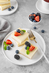 Vanilla cheesecake cut into squares with fresh berries on a white plate on a gray stone background