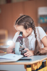 A cute girl, a first-grade elementary school student, looks through the eyepiece of a microscope at...