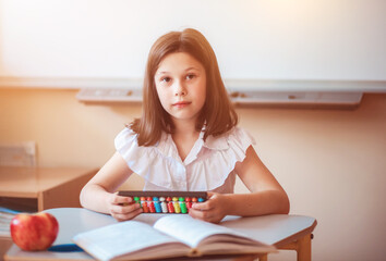 A cute girl sits at a school desk in the classroom and holds an abacus in her hands. A girl counts on an abacus during a math lesson.  Back to school.
