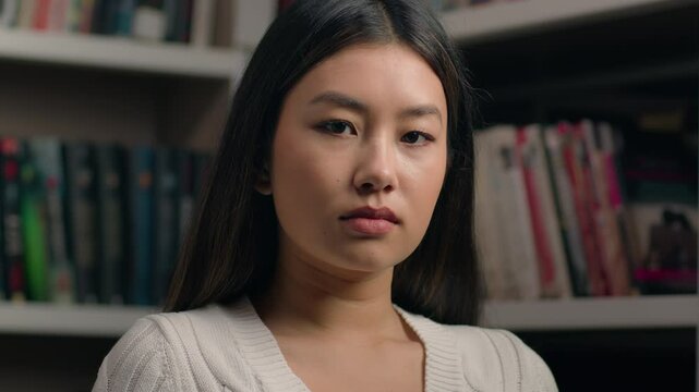 Serious concentrated asian woman girl student teenager businesswoman business lady teacher customer nod head approve agree looking at camera with piercing stern gaze posing indoors near bookshelf