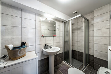 Standard modern bathroom featuring a corner shower cabin, sink, mirror, and light. The space is adorned with dark grey tiles, creating a sleek and contemporary look.