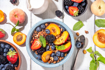 Granola breakfast with fruit and berry, summer whole grain tasty breakfast bowl with a lot of fruit and berries, with milk jug on white wooden table
