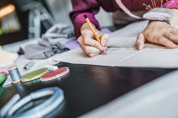 Cropped image of fashion designer woman working drawing her new design on a paper in her workshop. Female tailor hands draw sew pattern, ruler threads and needles on the table