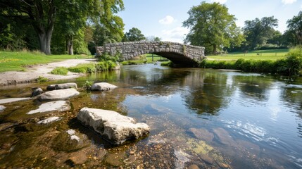 Fototapeta premium A charming stone bridge spanning across a clear, peaceful river with stones visible through the water and surrounded by lush greenery and trees under a bright blue sky.