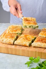 A rectangular Greek pie with spinach and cheese on a wooden board on a gray concrete background. Spinach recipes.