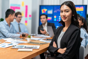 Portrait of happy young asian businesswoman or analyst looking at camera with her colleague analyzing data analysis in dynamic business strategy investment planning meeting. Habiliment