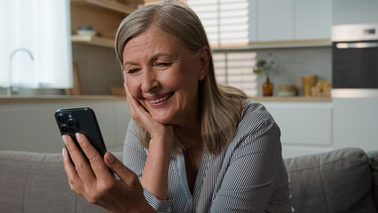 Carefree senior 50s happy Caucasian woman elderly grandparent smiling laugh sofa looking mobile phone smartphone mature female watch video online pensioner remote call with relatives use Internet home