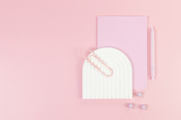 Top view of pink notebook, pink pen, paperclip, pins, white arch tray on pink background. School,...