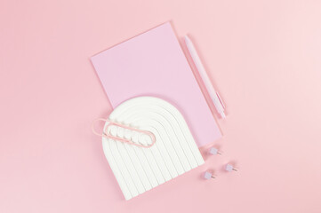 Top view of pink notebook, pink pen, paperclip, pins, white arch tray on pink background. School, office wallpaper. Flat lay, copy space.