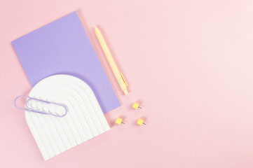 Top view of purple notebook, yellow pen, paperclip, pins, white arch tray on pink background....