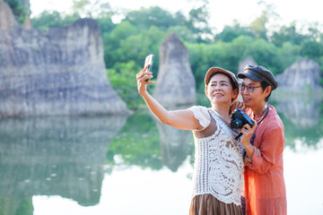 Two elderly Asian women, one in orange dress, other in white lace top, taking selfie by a river with green trees and rock formations in background. Smiling, enjoying friendship and nature.