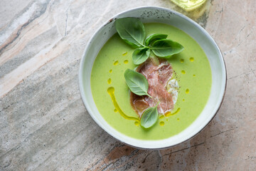 Bowl of green pea soup served with torn prosciutto and basil, horizontal shot on a grey and roseate granite background with space, high angle view