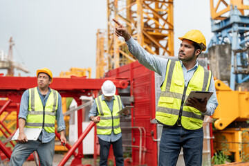Asian male engineer wearing safety vests and helmet with tablet pointing to tower crane. Team of...