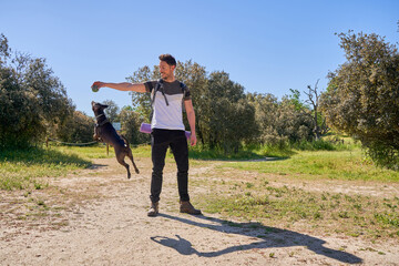 A man is playing with his dog in a park. The dog is jumping in the air to catch a ball. The man is wearing a white shirt and black pants.