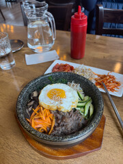Korean dish Bibimbap served in a restaurant in a hot stone bowl on a wooden table with metal chopsticks and a variety of banchan. There is a pitcher of water and the original sauce.