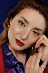 A stylish Asian woman in a red suit and silk shirt poses with a confident and alluring expression, her hands delicately framing her face.