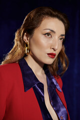 A stylish Asian woman poses in a red suit and silk shirt, with a confident expression and a hint of mystery.