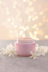 Cream in jar and beautiful jasmine flowers on grey table against beige background with blurred lights