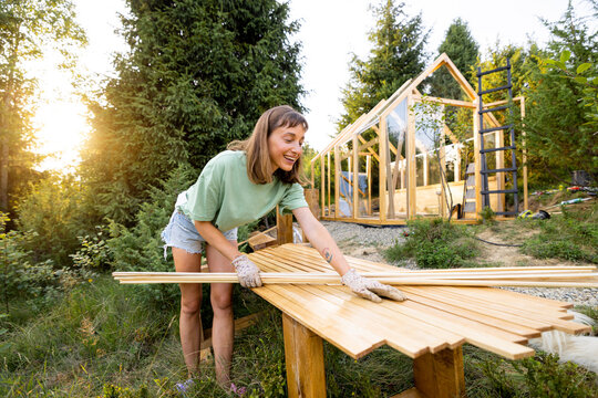 Smiling female carpenter holding wooden planks in front of a partially built greenhouse in her backyard. Engaged in a DIY project, surrounded by nature