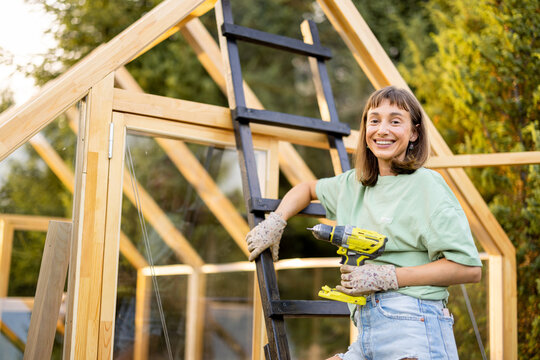 Smiling female carpenter holding a power drill next to a wooden ladder in front of a partially built greenhouse in her backyard. Engaged in a DIY project, surrounded by nature
