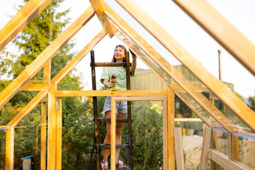 Female carpenter using a power drill while working on a wooden greenhouse in her backyard. She is...
