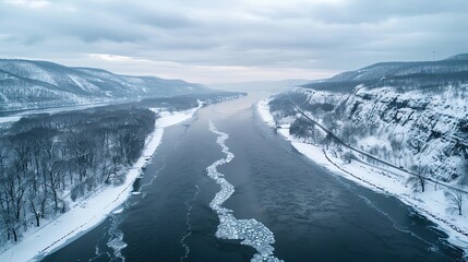 Aerial view of a frozen river cutting through snowy mountains and forests under a cloudy sky during winter.