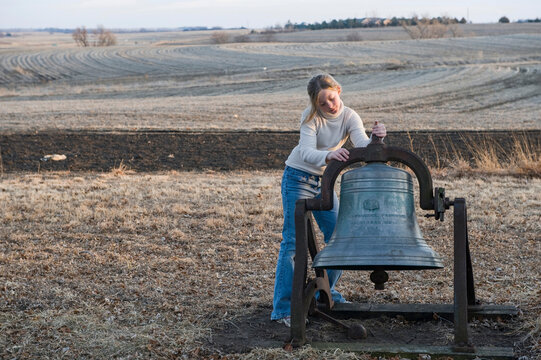 Girl stands by an old bell on farm in the vast counryside in Nebraska, USA; Cortland, Nebraska, United States of America