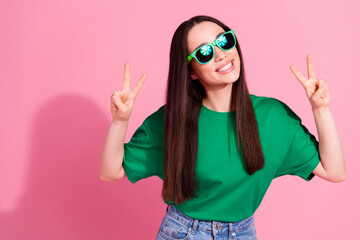 Photo of charming positive woman wear green t-shirt dark glasses showing two v-signs empty space isolated pink color background