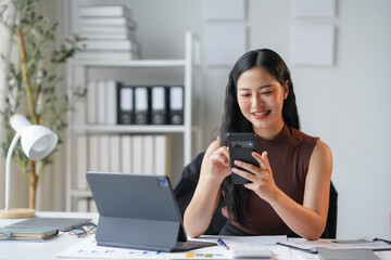 Fototapeta premium Young asian businesswoman multitasks happily in a modern office, using her smartphone and digital tablet at her desk