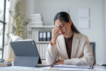 Stressed young businesswoman with a headache works on laptop in office, battling burnout and exhaustion from overwork and deadlines. Professional despite pain, analyzing finance documents