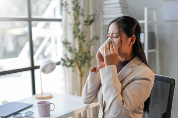 Businesswoman working in her office and blowing her nose with a tissue, she is sitting at her desk with a worried expression