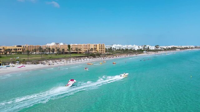 The image is an aerial view of a beach with crystal blue water of a Hotel at Mahdia, where people are enjoying various activities. A few surfboards can also be seen floating on the water.
