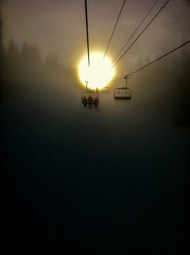 Ski chair lift with skiers going into snowy sun with pine trees