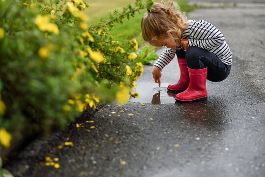 Toddler girl wearing rain boots pokes her finger into a puddle.