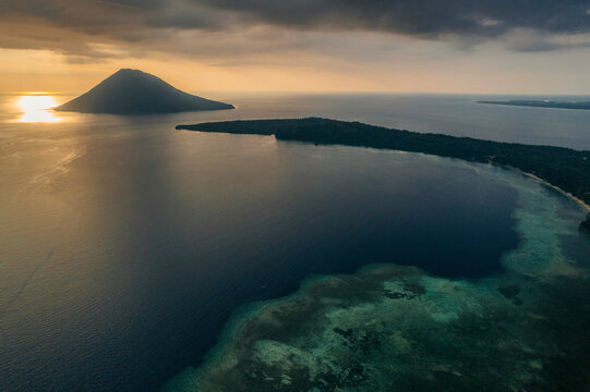 Drone view of Bunaken Island at sunset, with the warm sunlight reflected on the tranquil ocean and landforms silhouetted; Bunaken, North Sulawesi, Indonesia