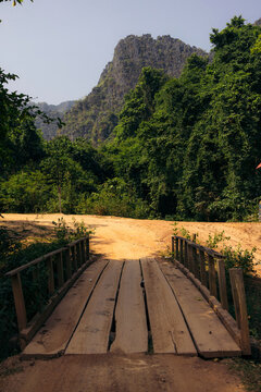 Wooden boards laid to cross water along a dirt road with lush vegetation and limestone karst formations against a blue sky; Ban Phahom, Luang Prabang Province, Laos