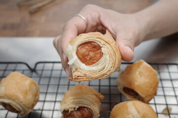 Hand holding a freshly baked sausage roll pastry with flaky golden layers, placed over a cooling rack. Close-up view of homemade bakery snack, warm and appetizing food concept.