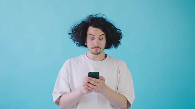 In front of a vibrant blue backdrop, a stylish young man with curly hair is deeply focused on his smartphone, symbolizing the impact of modern technology in contemporary lifestyle
