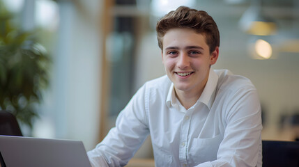 Obraz premium Portrait of a young male office worker, businessman smiling and posing at the camera and working on a laptop while sitting at a desk